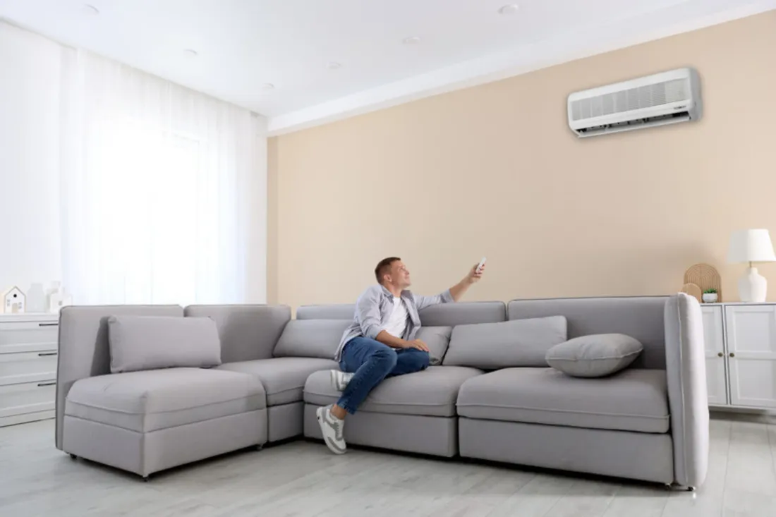 Mężczyzna na dużym szarym narożniku włącza pilotem klimatyzator na beżowej ścianie salonu