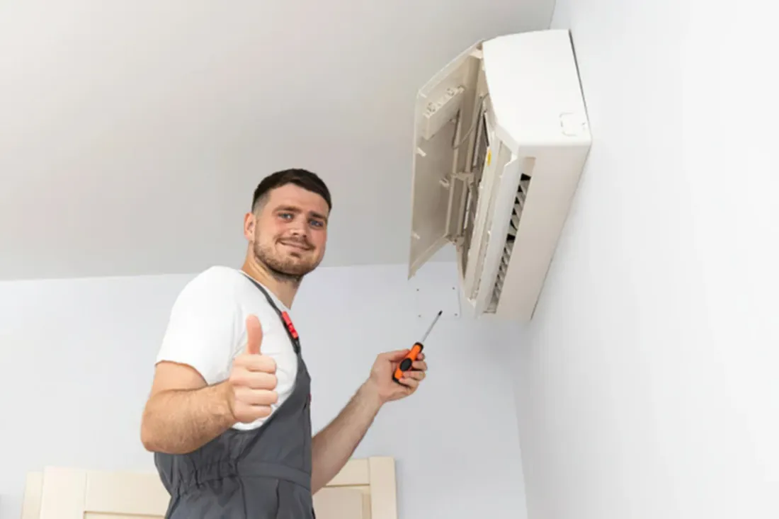 Uśmiechnięty monter ze śrubokrętem pokazuje kciuk w górę obok otwartego klimatyzatora