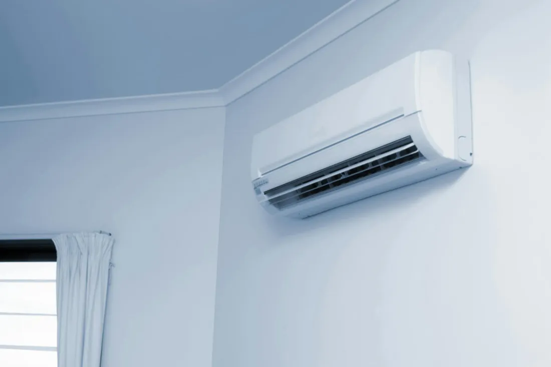 Klimatyzator ścienny tuż pod sufitem w narożniku jasnego pokoju, obok okno z zasłoną
