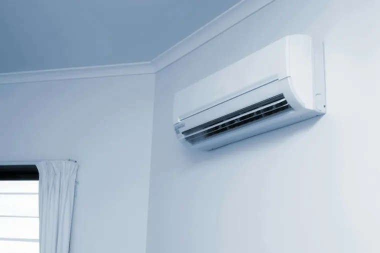 Klimatyzator ścienny tuż pod sufitem w narożniku jasnego pokoju, obok okno z zasłoną