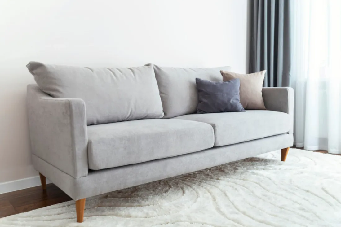 Jasnoszara dwuosobowa sofa na drewnianych nóżkach z granatową i beżową poduszką na puszystym białym dywanie