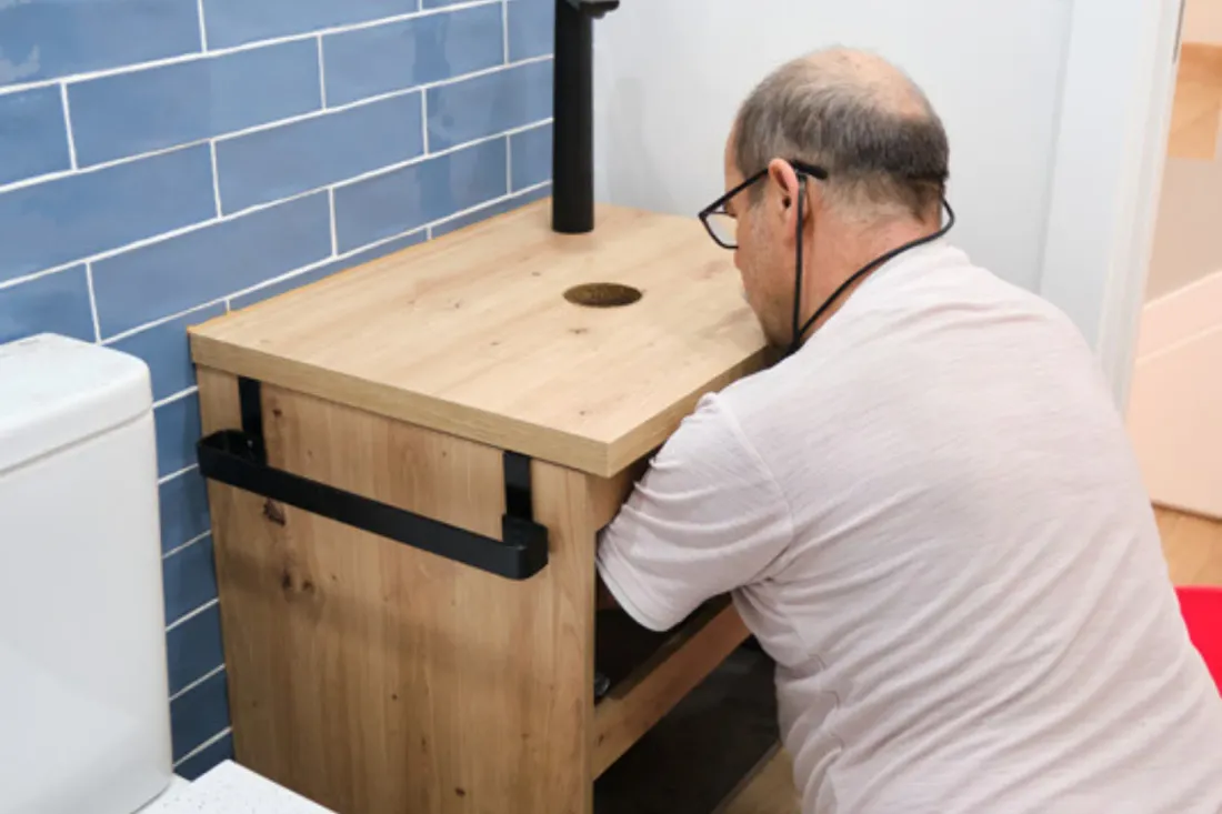 Starszy monter klęczy przy dębowej szafce podumywalkowej z czarną baterią i montuje odpływ w łazience z niebieskimi płytkami