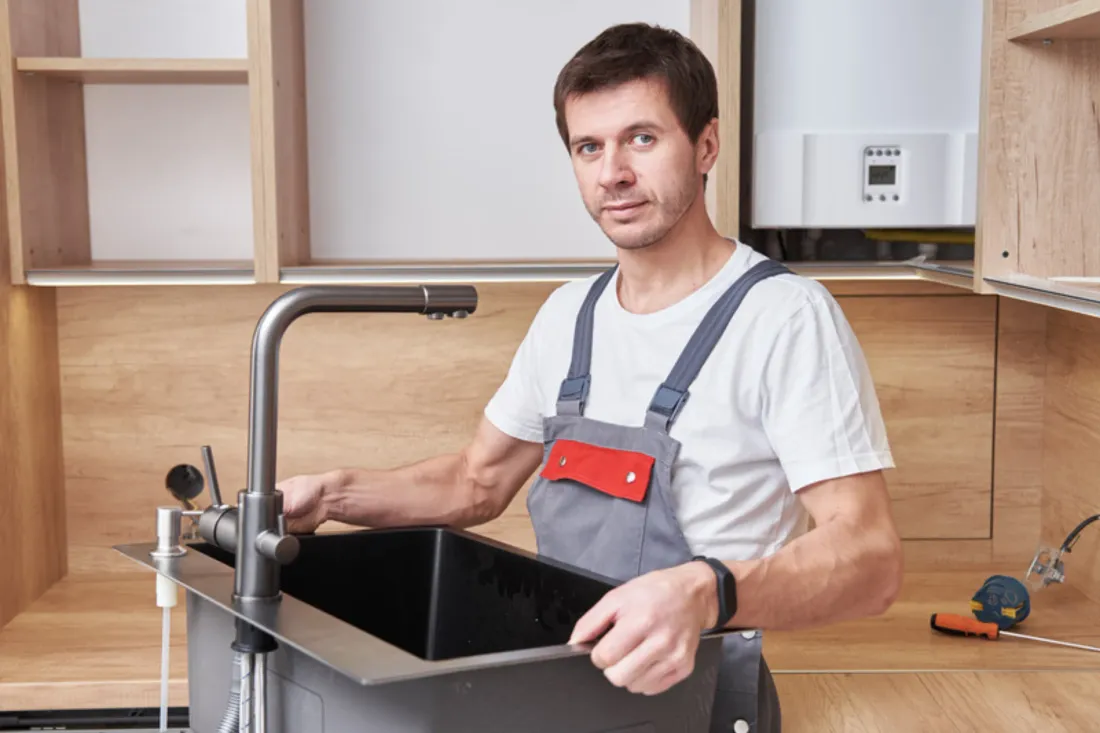 Hydraulik w szarych ogrodniczkach osadza czarny zlew z grafitową baterią w drewnianej zabudowie kuchennej